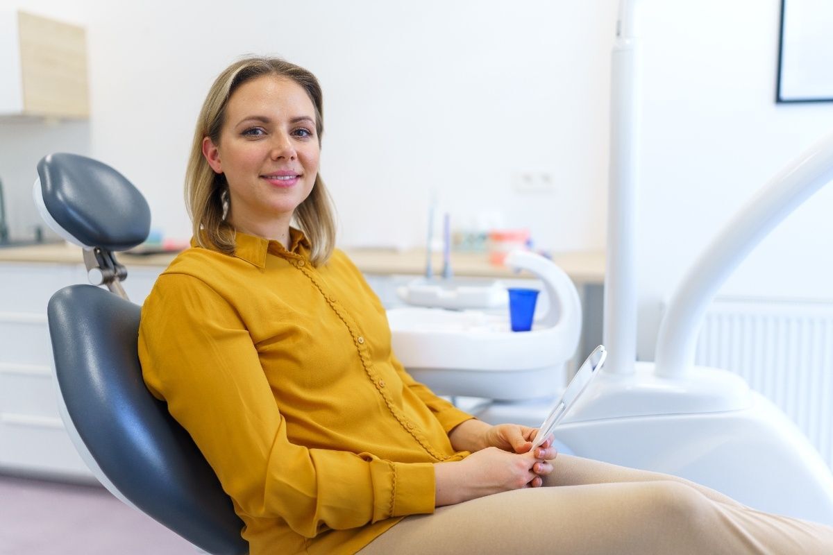 Woman in yellow shirt smiling while sitting in dental chair holding tablet in modern dental office.